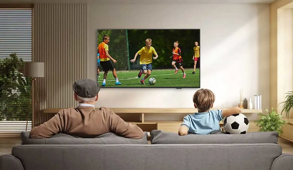 Een LG-tv hangt aan de muur in een moderne woonkamer, waar een man en jongen voetbal kijken; de jongen houdt een bal vast.