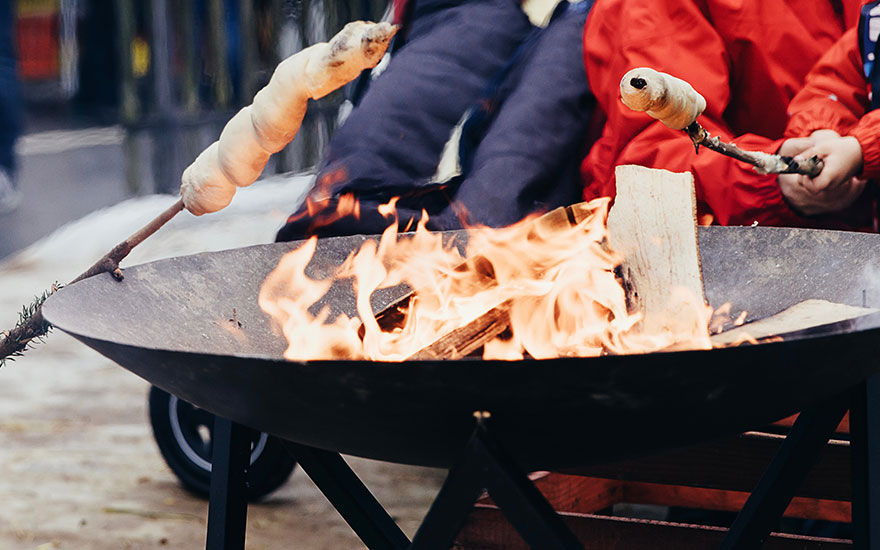 Open vuur in een metalen vuurkorf met brood op een stok boven de vlammen, klaar om geroosterd te worden.