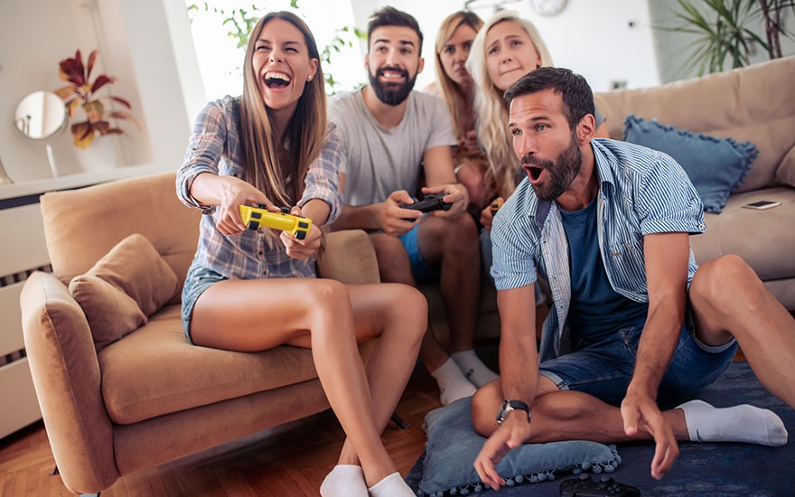 Vijf mensen zitten in een woonkamer met een beige sofa en spelen videospelletjes met controllers.