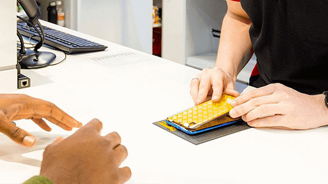 Un comptoir blanc avec un clavier, une souris et un téléphone jaune en réparation sur un tapis gris. Mains visibles.