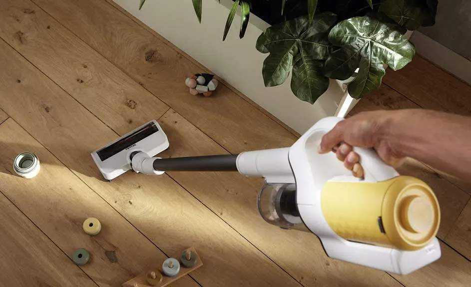 Un aspirateur jaune et blanc sur un plancher en bois, près d'une plante verte et de petits jouets en bois.