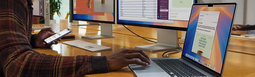 Persoon werkt aan een Apple MacBook met twee grote Apple-monitoren, gebruikt ook een iPhone; Apple-logo’s duidelijk zichtbaar.