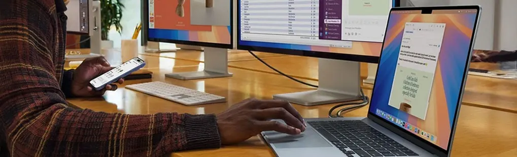 A person works on an Apple MacBook linked to two large monitors, while holding an iPhone, on a wooden desk with keyboards and tidy cables.