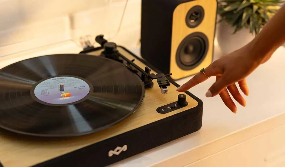 A hand operates a turntable with a black vinyl record. A yellow speaker stands nearby. Warm, rustic setting with green plant decoration on a wooden surface.