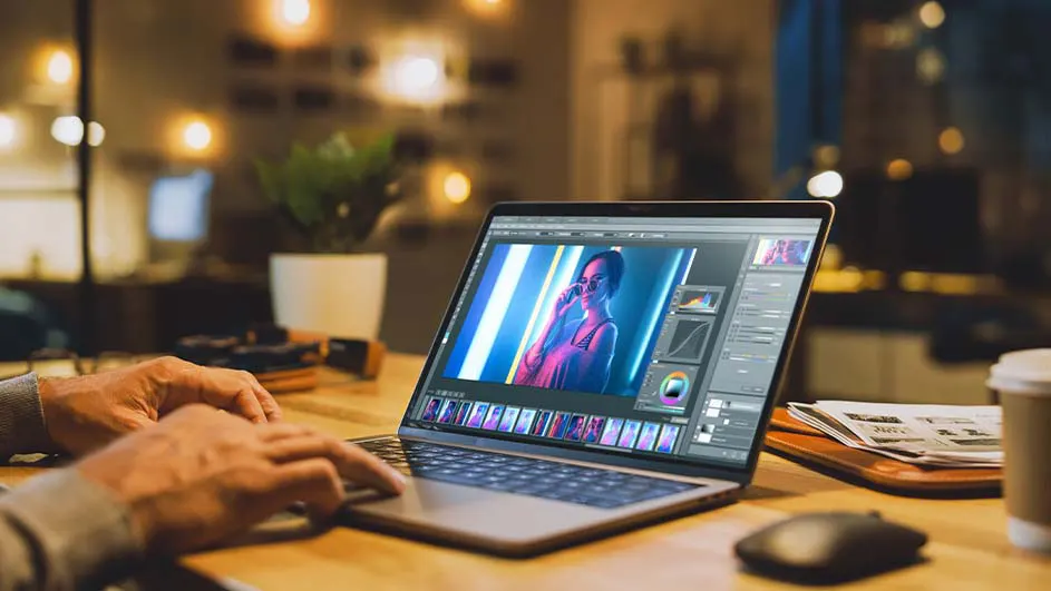 Een laptop staat open op een houten bureau met handen die typen. Op het scherm is een foto te zien. Een plant, kop koffie, en muis staan op tafel.