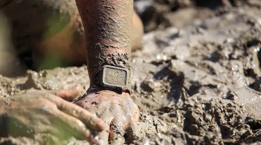 Une main et un bras couverts de boue, avec une montre carrée au poignet, sur un fond boueux.