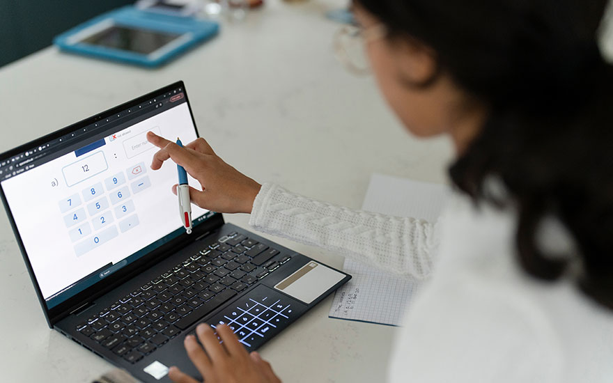 Een zwarte laptop toont een rekenmachine op het scherm, bediend met een pen. Een blauwe tablet ligt op een witte tafel.