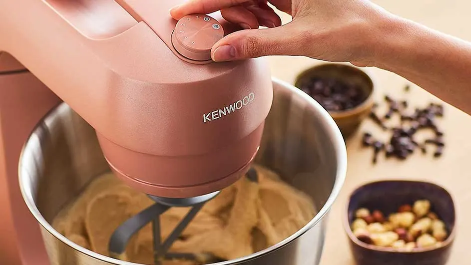 Een roze Kenwood mixer mengt deeg in een metalen kom. Schalen met chocolade en noten staan op een lichtbruine tafel.