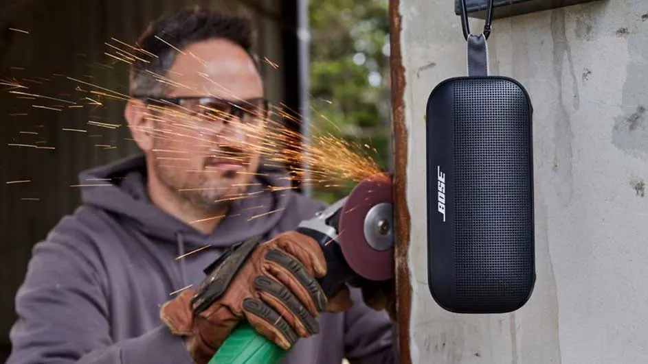 Un homme utilise une meuleuse, produisant des étincelles, près d'une enceinte Bose noire suspendue à un mur.