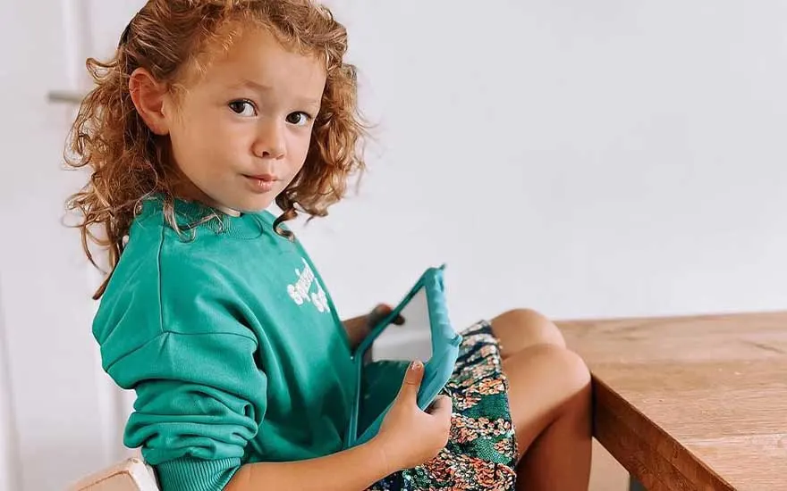 Une jeune enfant en pull vert tient une tablette protégée et est assise à table, prête à jouer ou à apprendre.