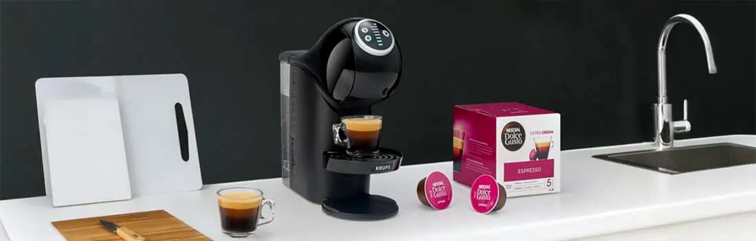 Een zwarte Dolce Gusto-koffiemachine zet espresso op een wit aanrecht, naast roze capsules, een doos, snijplanken en een gootsteen.