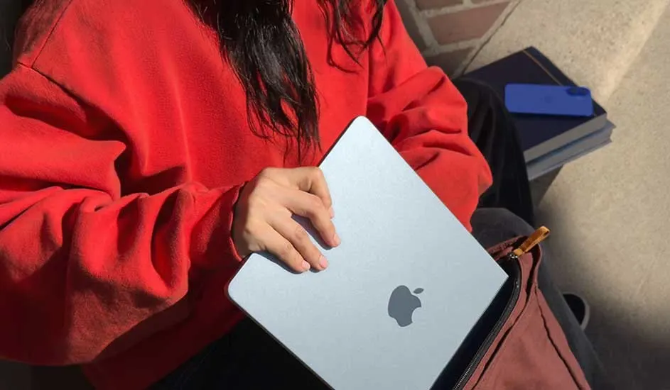 Persoon in rode trui stopt een zilveren MacBook in een tas, naast haar liggen boeken met een blauw hoesje op zonnige stenen.