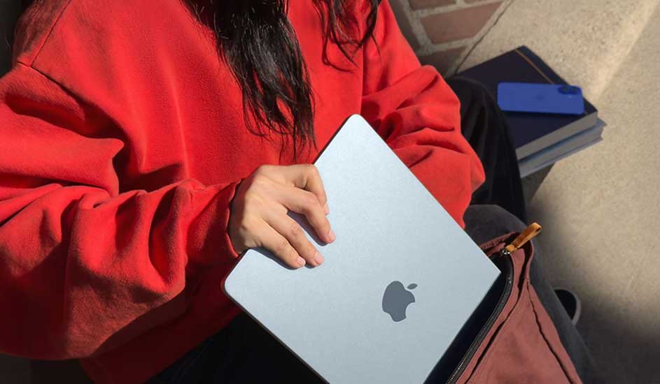 Person in a red sweatshirt putting a silver MacBook into a bag, with books and a blue phone case nearby on sunlit steps.