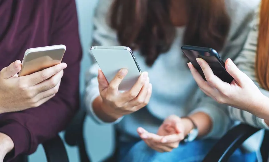 Drie mensen zitten op stoelen en kijken naar hun smartphones. De telefoons zijn goud, zilver en zwart.