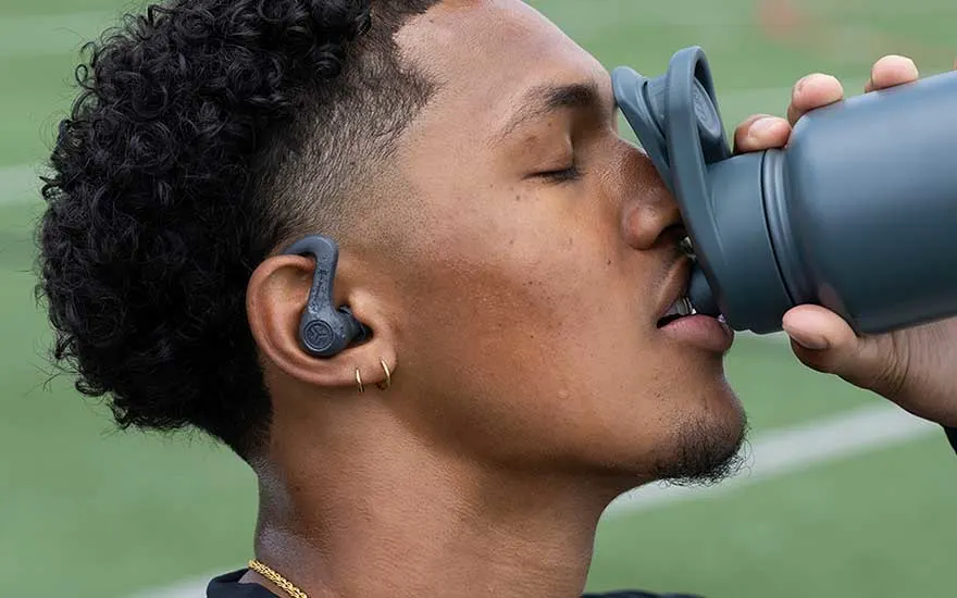 Person with wireless earbuds drinks from a gray water bottle while exercising on a grass field.