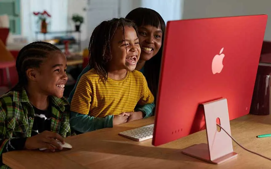 Twee kinderen en een volwassene kijken lachend naar een rode Apple iMac op een houten bureau met een toetsenbord en een muis.