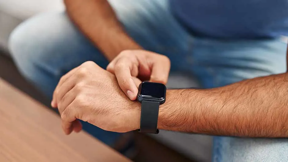 Gros plan sur une montre noire à écran tactile au poignet, avec un bracelet noir. Le fond est bleu et bois.