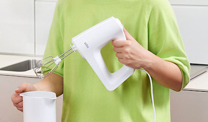 Iemand houdt een witte mixer boven een witte maatbeker in een lichte keuken.