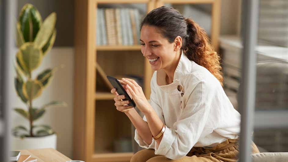 Une personne utilise un smartphone. Une plante verte et une bibliothèque sont visibles en arrière-plan.