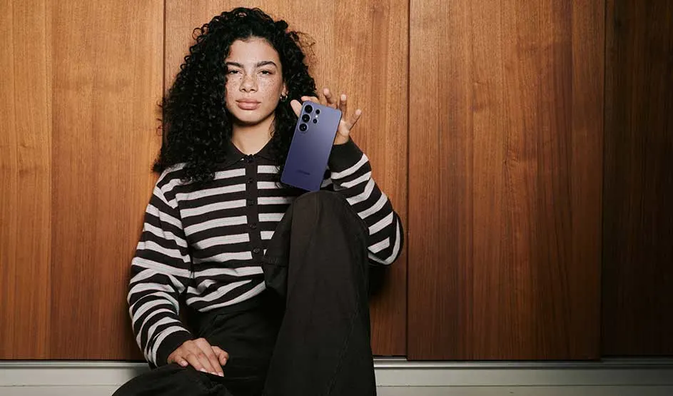 Jongvolwassene zit relaxed tegen een houten wand en toont een donkerblauwe smartphone, het toestel duidelijk in de spotlight van Samsung!