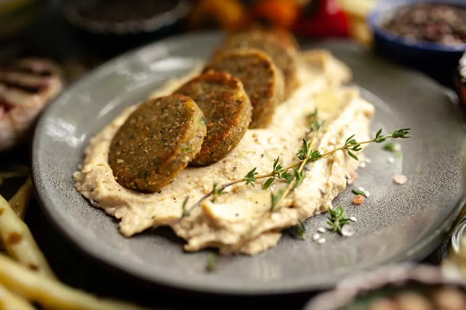 Vier bruine falafel op een bedje van beige hummus op een grijs bord, afgewerkt met een takje groene tijm.