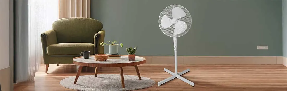 Un ventilateur sur pied blanc se trouve dans un salon moderne, à côté d’un fauteuil vert, d’une table en bois et de plantes.