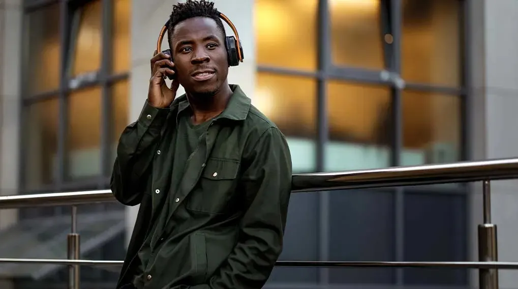 Een man staat buiten met een donkergroen hemd en een zwarte koptelefoon tegen een gebouw met ramen.