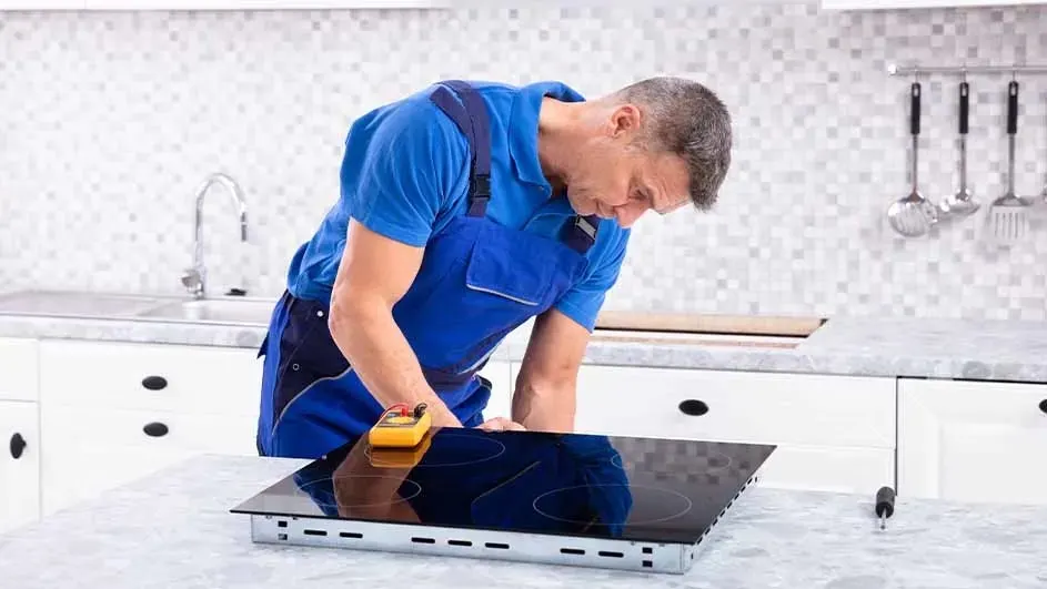 Un homme en bleu répare une plaque de cuisson noire dans une cuisine blanche. Un multimètre jaune est visible.