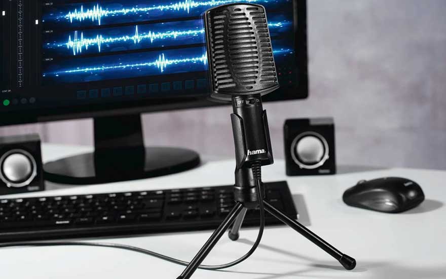 Un microphone noir sur un trépied devant un écran d'ordinateur affichant des ondes sonores bleues, avec un clavier, des haut-parleurs et une souris.