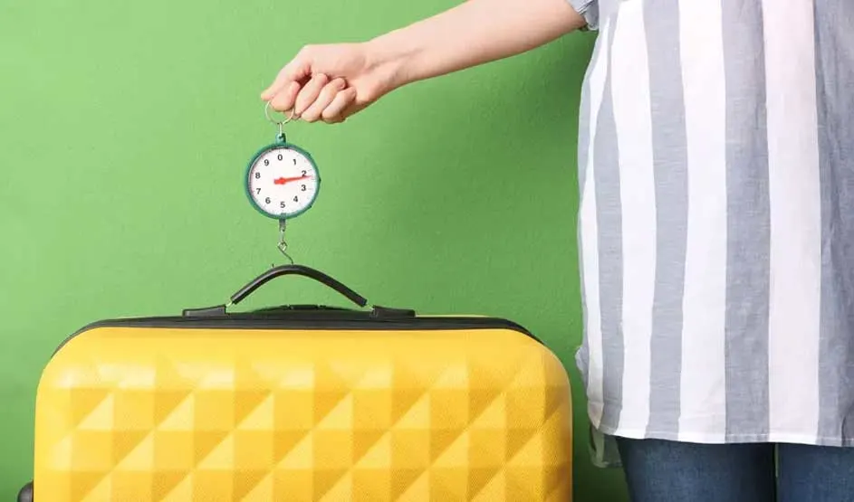 Une main soulève une valise jaune à l'aide d'une balance sur un fond vert uni. La valise a une poignée noire.