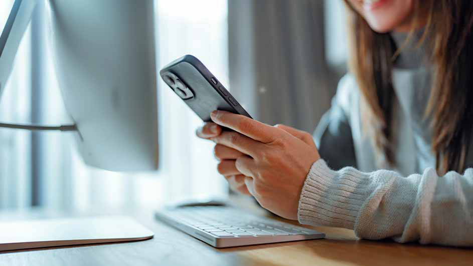 Une personne utilise un smartphone noir devant un écran d'ordinateur et un clavier blancs.