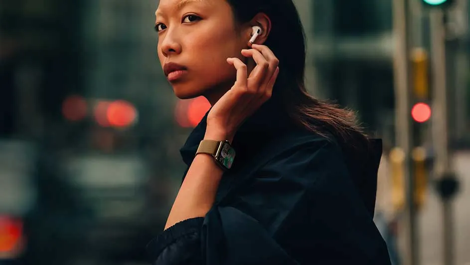 Een close-up van een horloge met een beige band, een witte oortelefoon en lichtreclame op een donkere achtergrond.