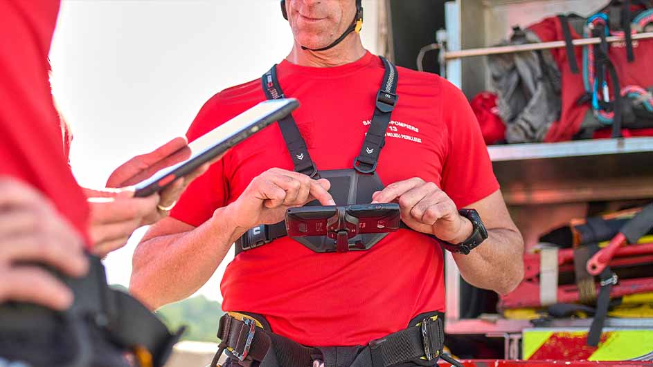 Een brandweerman in rood uniform bedient apparatuur op zijn borst, met een tablet in de hand en een brandweerwagen op de achtergrond.