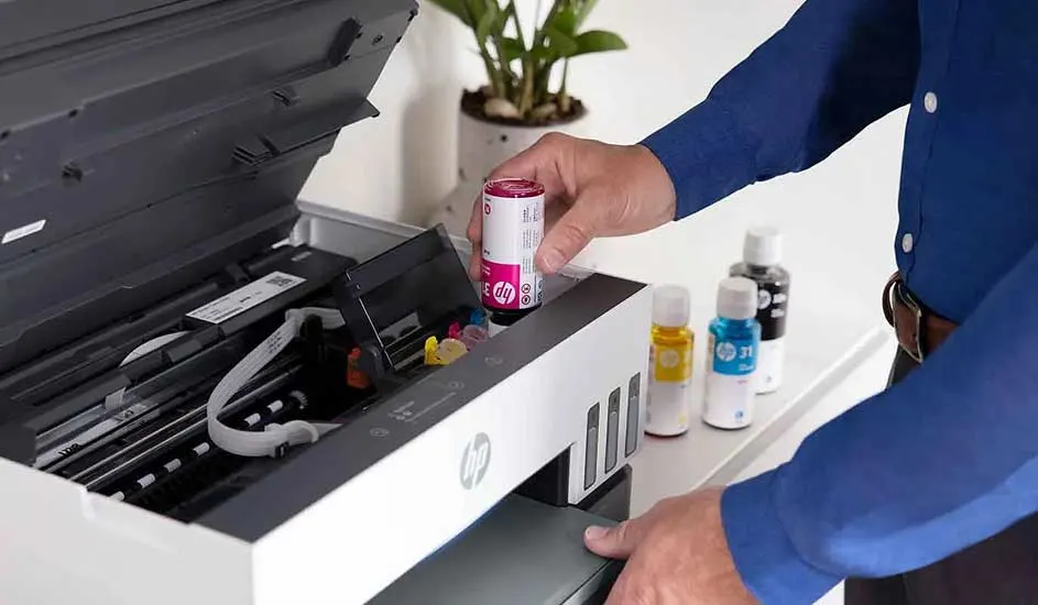 Een printer wordt bijgevuld met inkt. Er staan flessen inkt (geel, cyaan, zwart) naast de printer. De printer is wit en grijs.