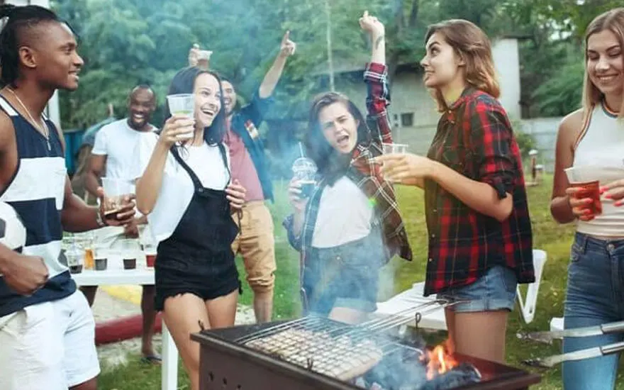 Groupe de personnes à un barbecue dans un jardin. Un gril est au premier plan, avec de la fumée et des flammes visibles.