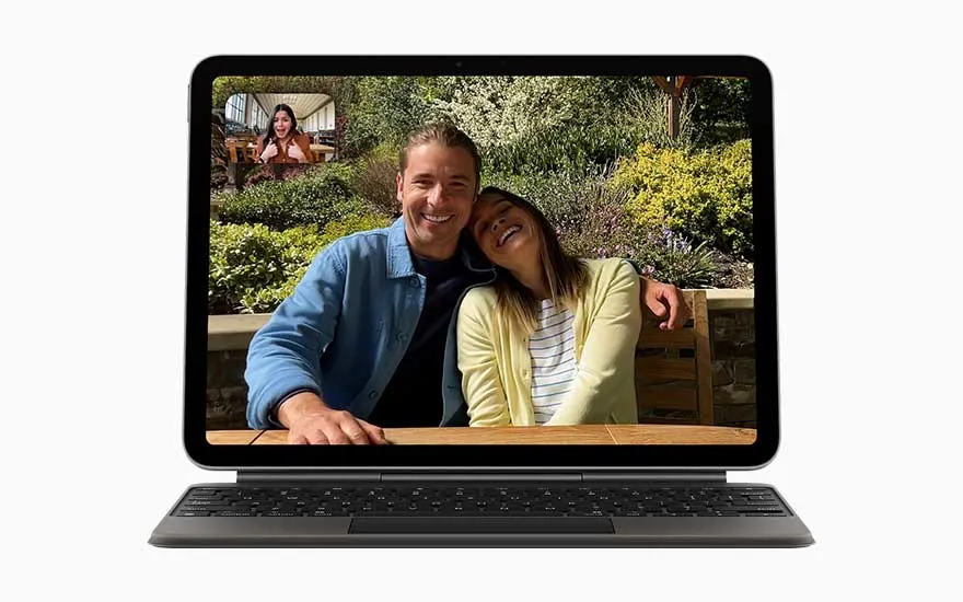 Un iPad avec clavier noir affiche un appel vidéo : un couple souriant est assis dehors, tandis qu’une personne apparaît en haut à gauche.