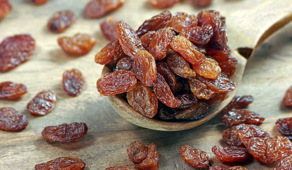 Un tas de raisins secs bruns dans une cuillère en bois, éparpillés sur une table en bois.
