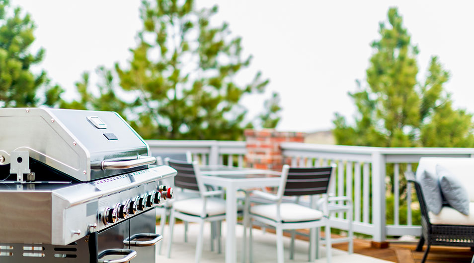 Een zilveren barbecue staat op een terras met witte meubels en groene bomen op de achtergrond.