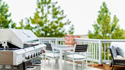 Un barbecue en acier inoxydable sur une terrasse avec table, chaises blanches et arbres verts en arrière-plan.