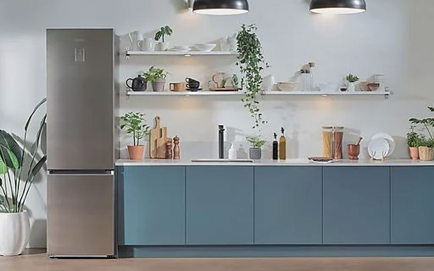 Cuisine avec frigo gris, armoires bleues, étagères blanches avec plantes et vaisselle, sous deux lampes suspendues.