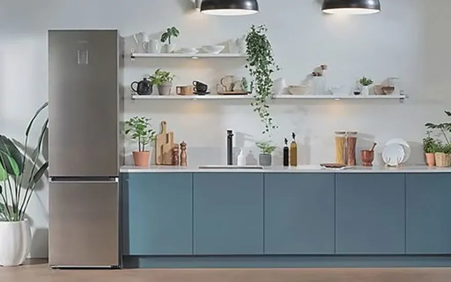 Cuisine avec frigo gris, armoires bleues, étagères blanches avec plantes et vaisselle, sous deux lampes suspendues.