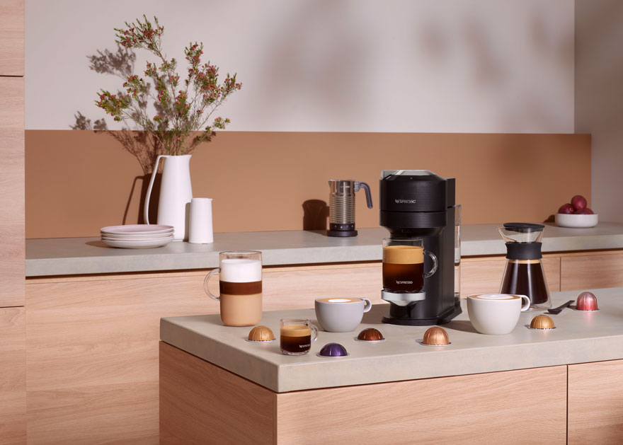 Une machine à café noire prépare du café dans une tasse transparente. Des tasses, des capsules et des fleurs décorent le comptoir.