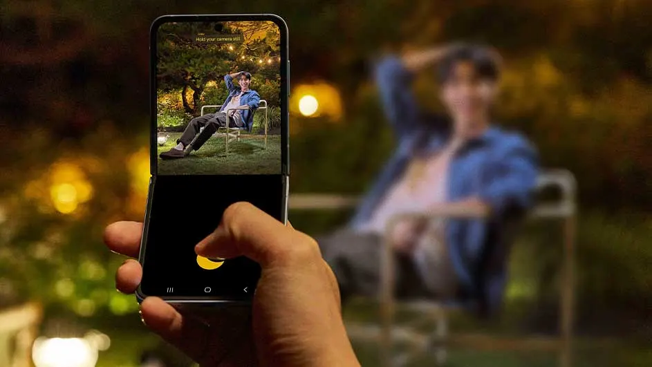 Main tenant un smartphone pliable, un doigt appuie sur l'écran. L'écran supérieur affiche une personne assise dans un jardin éclairé.