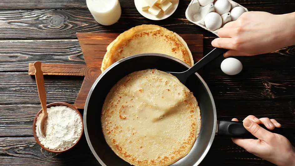 Crêpes en poêle, lait, œufs et farine sur table en bois. Une spatule noire retourne la crêpe.