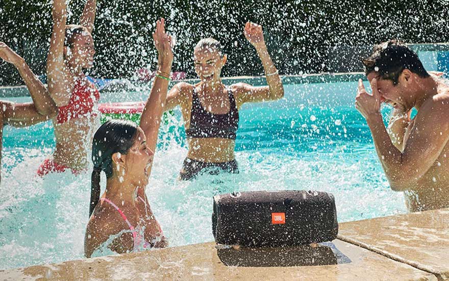 Haut-parleur noir JBL sur une terrasse beige. Arrière-plan : personnes dans une piscine bleue avec des éclaboussures d’eau.