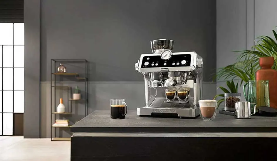 Une machine à café chromée sur un comptoir gris, avec des tasses de café, devant un mur gris et des plantes.