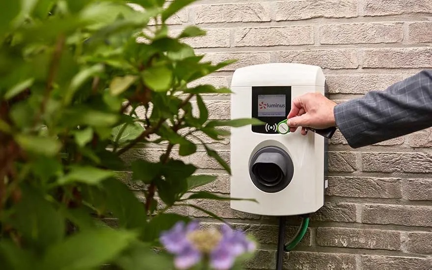 Close-up of a white EV charger mounted on a brick wall, activated with a green key fob. Green leafy plants surround the charger.