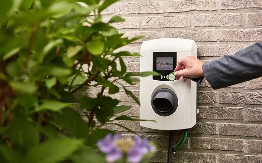 Close-up of a white EV charger mounted on a brick wall, activated with a green key fob. Green leafy plants surround the charger.