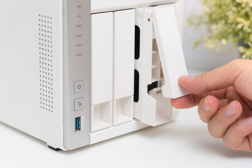 Un serveur NAS blanc est manipulé par une main. Plusieurs baies de stockage sont visibles. Un port USB est visible à l'avant.