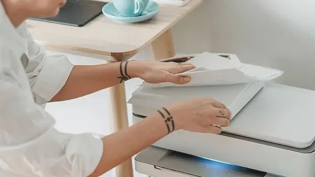 A person places papers on an HP printer or scanner next to a desk with a cup and a laptop.